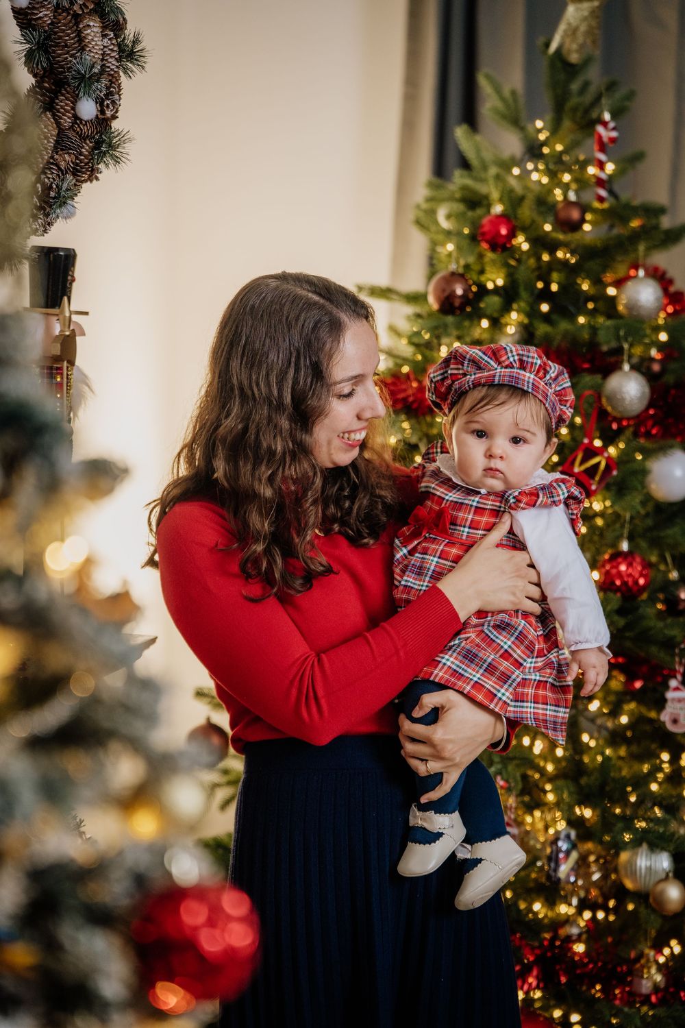 Mãe sorridente com a filha bebé ao colo, de vestido tartan, junto à árvore de Natal