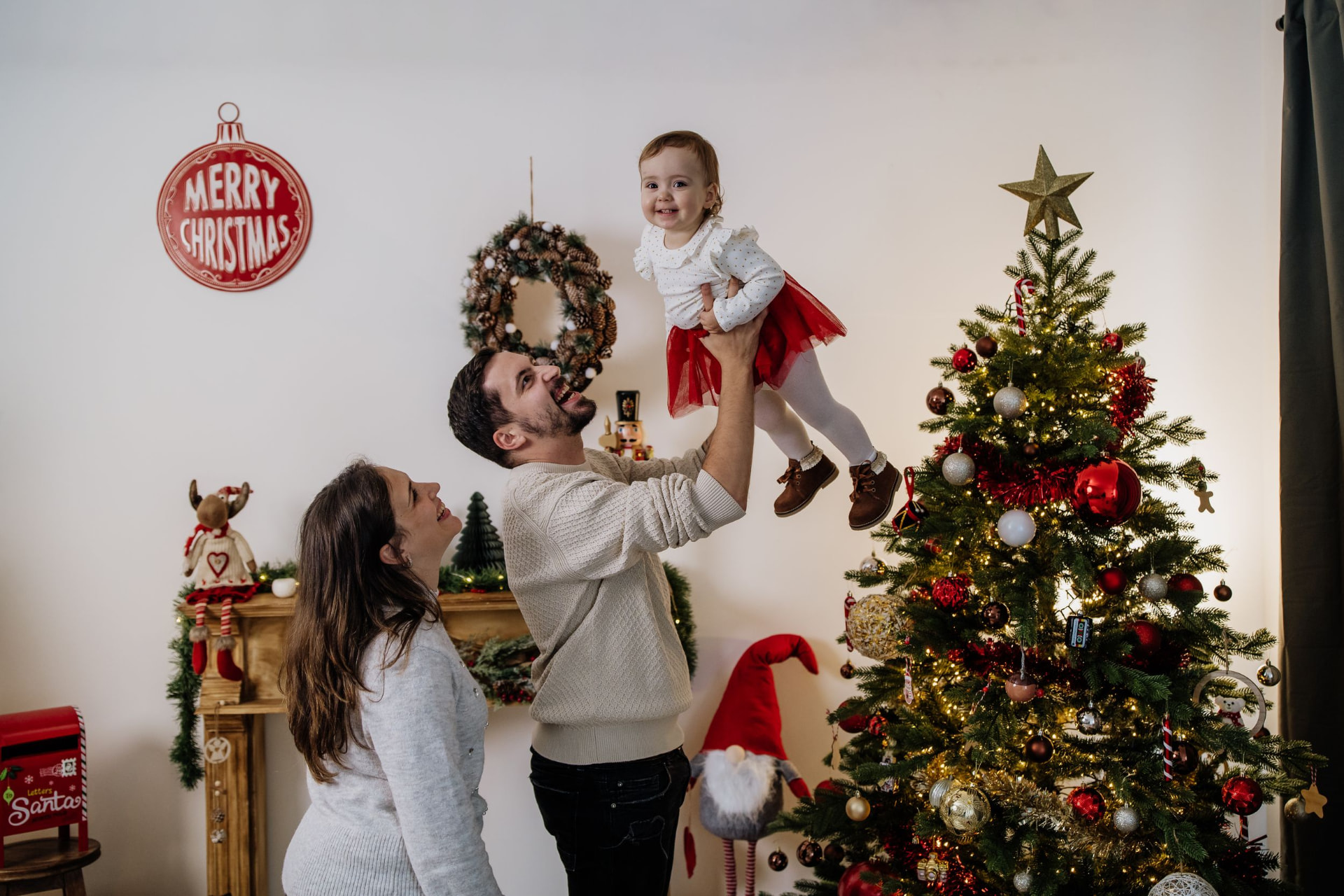 Pais a levantar a filha bebé ao colo, junto à árvore de Natal iluminada