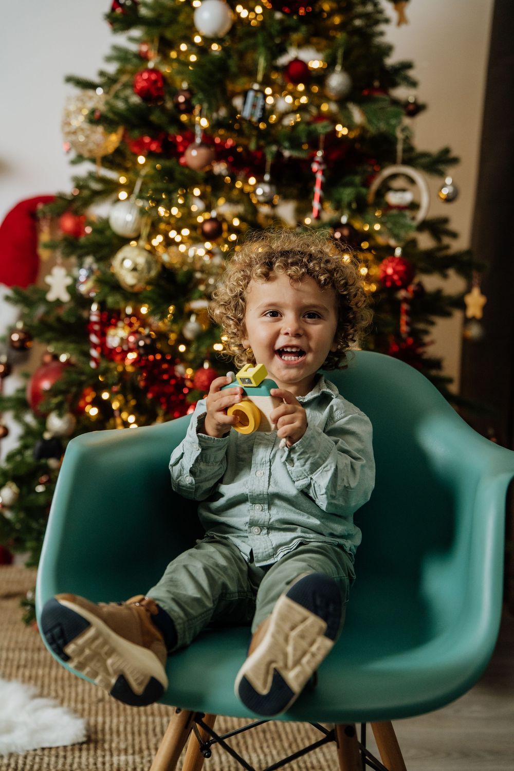 Criança sorridente com uma câmara de brincar, sentada numa cadeira verde junto à árvore de Natal