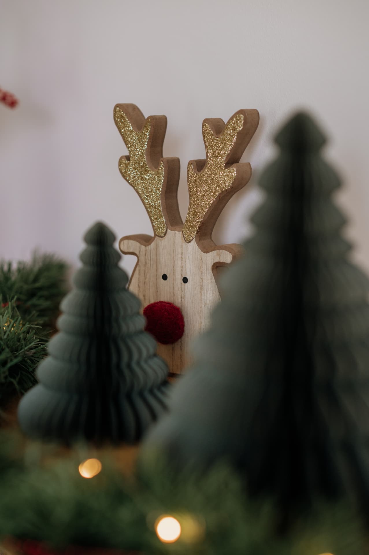 Detalhe de decoração de Natal: rena de madeira com chifres de purpurina dourada, entre árvores de papel verdes e luzes quentes